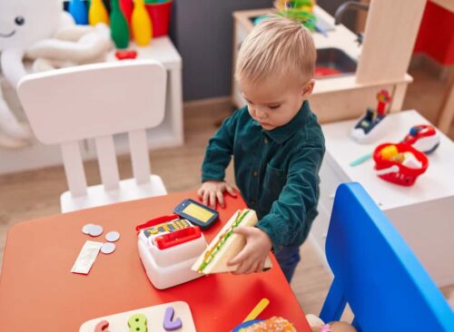 Adorable Caucasian Boy Holding Sandwich And Supermarket Toy At Kindergarten